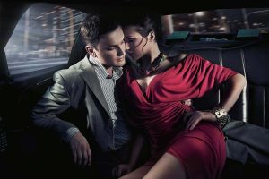 man in a suit and woman in a red dress cuddling together in a taxi while she smells his pheromones